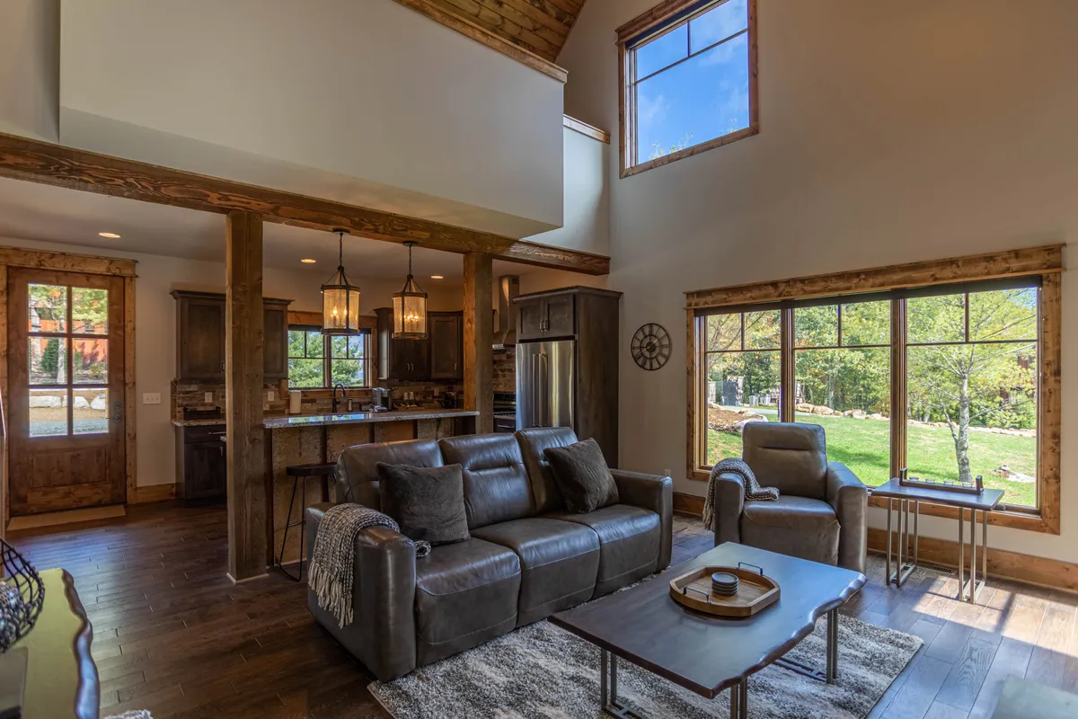 A spacious interior with large windows, abundant natural light, plush seating, and exposed wooden beams. The kitchen area is visible in the background, suggesting the loft’s connectivity to other living spaces. The scene exudes warmth and comfort, inviting guests to unwind or gather for conversation.