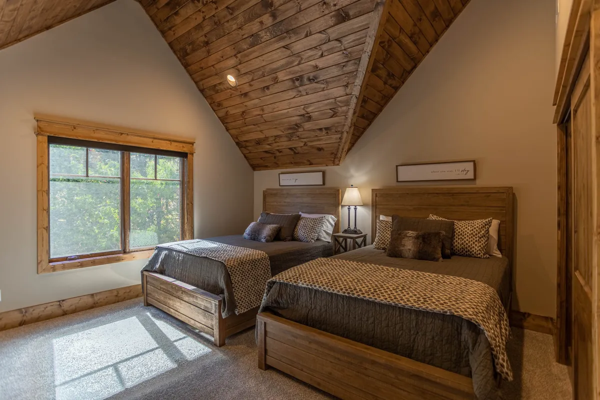 A cozy upstairs bedroom with two beds, a wooden ceiling, a large window, and a lamp. The space is bathed in soft natural light, evoking warmth and togetherness—well-suited for families or groups.