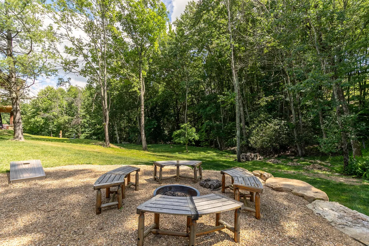 The image shows a fire pit area ringed by wooden benches, surrounded by mature trees. The setting is perfect for relaxing evenings, storytelling, and stargazing under clear mountain skies.