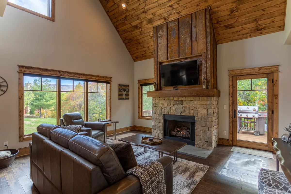 A warm, welcoming living room anchored by a stone fireplace, with comfortable seating, large windows offering natural light, and exposed wooden beams overhead. The room is arranged for both relaxation and socializing, creating an ideal spot for movie nights or quiet evenings by the fire.