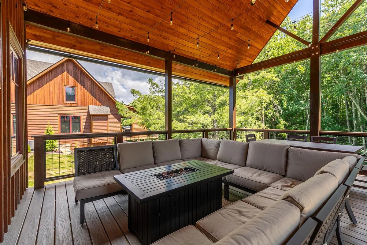 A welcoming outdoor seating area on a private deck, featuring a comfortable sectional, fire pit table, and wooden beams overhead. Lush greenery surrounds the deck, creating a tranquil atmosphere ideal for relaxing, dining, or stargazing.