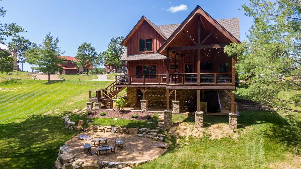 The image showcases a charming, newly built two-story cabin nestled in the lush hillside of Banner Elk's Eagles Nest community. The exterior features a spacious deck perfect for morning coffee, a beautifully landscaped lawn, and a cozy fire pit area set against mountain views, inviting guests to gather under the stars.
