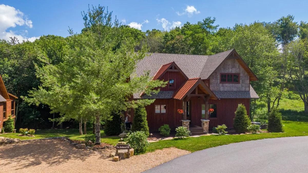 A charming red exterior of The Sheltered Elk stands out, surrounded by a landscaped front yard and decorative flower pots. The inviting porch features comfortable seating, suggesting a space where families and groups can relax outdoors and enjoy the natural setting. The warm lighting and subtle seasonal decor evoke a sense of welcome and togetherness.
