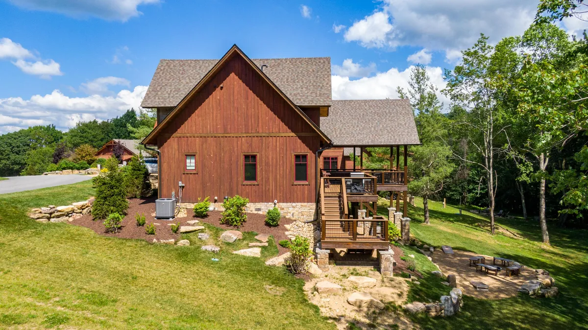 A picturesque exterior shot showing a wooden house with a spacious deck area, landscaped garden, and a cozy sitting area outlined with stones. The deck invites guests to gather for outdoor meals, relax in serene surroundings, or enjoy evenings by the fire pit. The scene is nestled in natural beauty, emphasizing the tranquil mountain retreat ambiance.