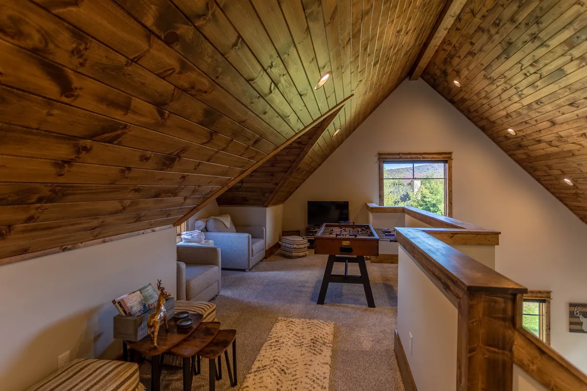 A comfortable loft area featuring a game table, seating, exposed wooden beams, and a window view. The setting is inviting for quiet play, relaxation, or reading, providing additional private space for guests.